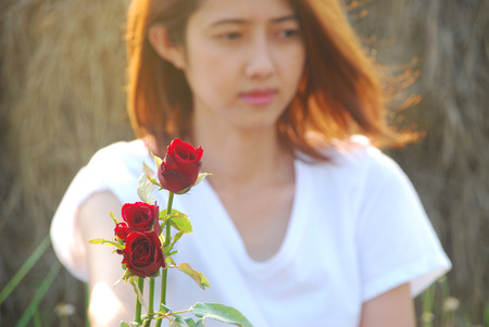 Asian young woman are moment in love are blurred background. She sit on the field in the evening sunshine have a mild breeze and holding a red rose in hand.の写真素材