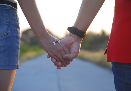 Lovers walk and holding hands on the road in evening. They have the necklace in heart shape is mean...
