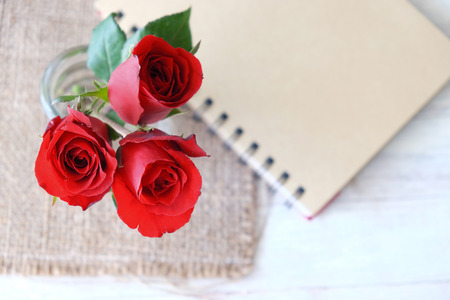Three red rose in a vase in the top view on sackcloth and notebook in blurred background.の写真素材