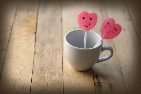 Pink heart shaped lollipops in lover concept have a happy face and decorated in a small white cup of coffee. Placed on a wooden floor, Adjust photo in retro styleの写真素材