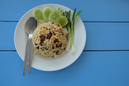 Fried rice with Chinese sausage in a white dish. Decorated with onion, cucumber and lemon slice with spoon and fork lay on blue wooden table in top view.の写真素材