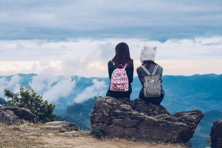 Two girls sitting back  on a rock  looking each other ,Blue sky with clouds and green trees beautiful view from aboveの写真素材