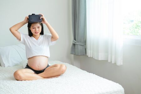 Pregnant Asian woman is wearing a Virtual Reality device sitting in a bed in the bedroom.の写真素材