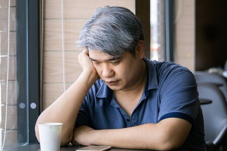 Stressed tired young Asian middle-aged man,old man take hand on head feeling depression and exhausted sitting by the window at coffee shop,grey hair man ,Family issues.の写真素材