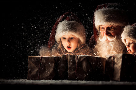 A magical winter scene featuring joyful children and Santa Claus as they discover opened giftboxes amidst falling snow.の素材