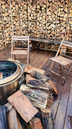 Cozy outdoor patio with stacked firewood wall, metal fire pit, and rustic wooden chairs on a wet...