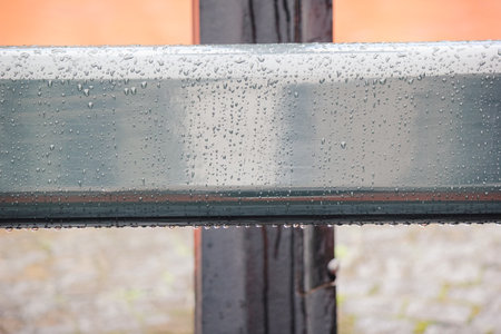 Close up of a smooth, gray metal railing covered in fresh rain drops with a blurred orange...