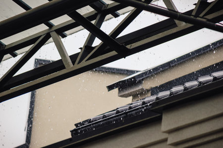 Heavy rain pouring off a black tiled roof and metal gutter below a modern, transparent canopy structureの写真素材