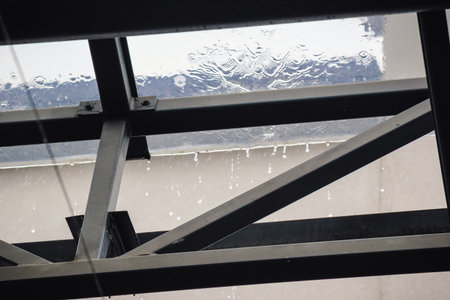 Rain falling on a transparent glass roof with water dripping from the dark metal support structure belowの写真素材