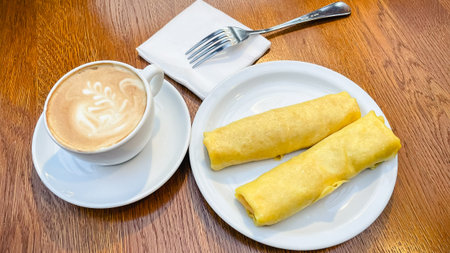 Delicious rolled pancakes served on a plate with a cup of hot cappuccino on a wooden cafe tableの写真素材
