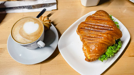Freshly baked savory croissant with lettuce served on a plate next to a cup of hot cappuccino on a cafe tableの写真素材
