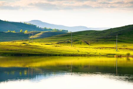 Green hills with power lines and their reflections in the lake. Beautiful summer landscape. South Ural, Russiaの写真素材