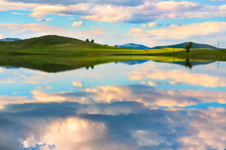 Green hills, blue sky with clouds and their reflections in the lake at sunset. Beautiful summer...