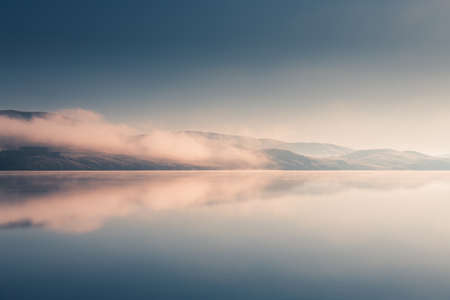 Beautiful lake in misty morning. Mountains and clouds are reflected in the calm water surface....