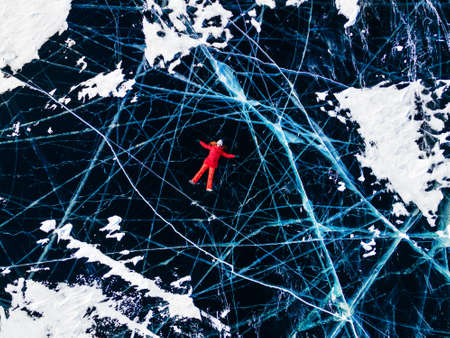Girl in red jacket lying on the ransparent cracked blue ice on Baikal lake. Aerial drone view. Baikal, Siberia, Russia. Winter travel destinationsの写真素材