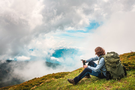 Young man traveler with backpack sitting on the top of the mountain with clouds and enjoying the view. Summer landscape. Travel and adventure conceptの写真素材