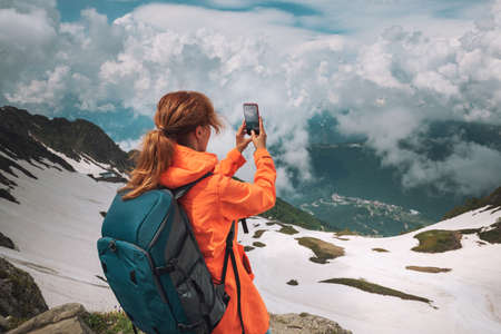 Young woman traveler with backpack standing on the top of the mountain and taking photo with smartphone. Summer landscape of the mountains and sky with clouds. Travel and adventure conceptの写真素材