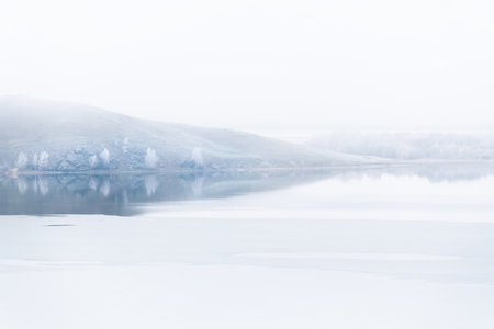 Mountains and frosted trees are reflected in the lake. First ice on the lake. Beautiful winter landscape at foggy morningの写真素材