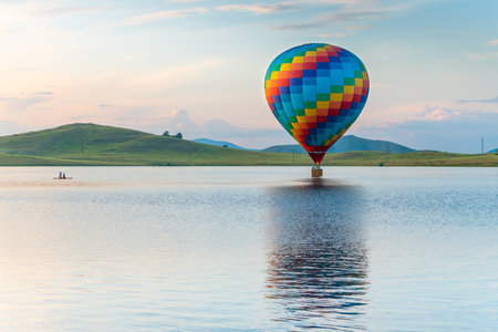 Colorful hot air balloon flying over the lake at sunset. Green hills and the blue sky in the background. Beautiful summer landscape.の写真素材
