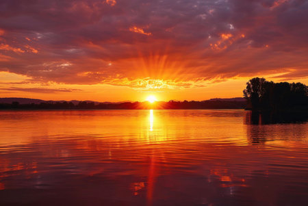 Stunning sunset over a tranquil lake. The sky is ablaze with vibrant orange and red hues, reflecting beautifully on the water.の写真素材