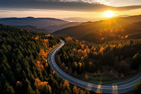 A winding road snakes through a vibrant autumn forest, bathed in the warm glow of a setting sun. Mountains and a peaceful lake are seen in the background.の写真素材