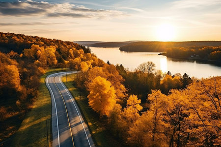 A scenic autumn road winds through vibrant golden trees, overlooking a tranquil lake at sunset. Breathtaking nature view.の写真素材