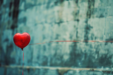 A single red heart-shaped balloon floats against a blue and green textured backdrop. The simple composition and vibrant color create a sense of love and optimism.の写真素材