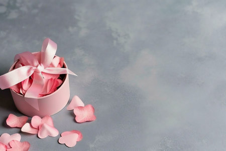 Pink gift box with a delicate ribbon tied on top. The box is filled with pink rose petals and surrounded by other scattered petals on a gray background.の写真素材