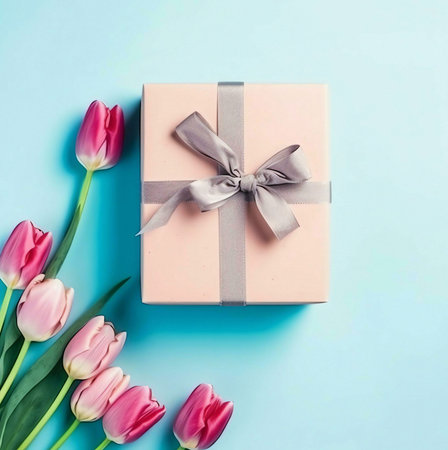 A pink gift box with a silver ribbon and bow on a blue background, surrounded by pink tulips.の写真素材