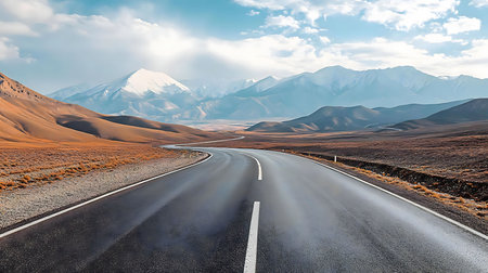 A scenic road cuts through a vast landscape, leading towards majestic snow-capped mountains under a brilliant sky.の写真素材