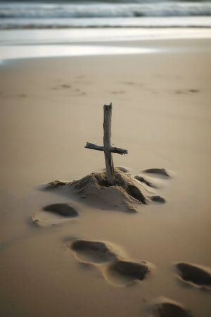 A simple wooden cross planted in the sand of a beach, with the ocean in the background. Footprints around the cross add to the contemplative mood.の写真素材