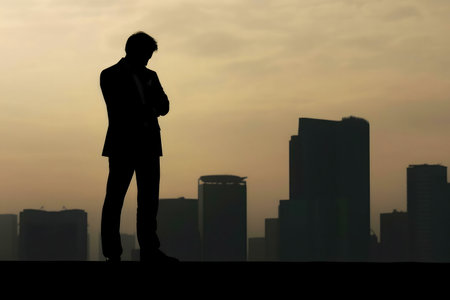 A pensive businessman stands silhouetted against a city's skyline at sunset. The image evokes a sense of contemplation.の写真素材