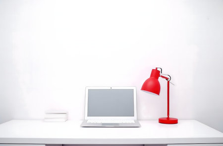 A clean white desk with a laptop, a red lamp, and some books. Minimalist workspace.の写真素材