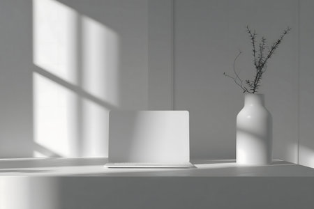 A serene minimalist workspace featuring a white laptop, a vase with branches, and sunlight casting shadows.の写真素材