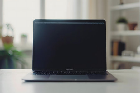 A sleek black laptop sits on a white desk in a bright, modern room. The screen is dark, and the background is softly out of focus.の写真素材