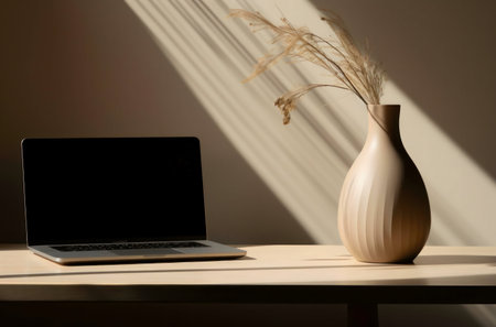 A laptop and a vase with dried flowers sit on a desk bathed in sunlight.の写真素材