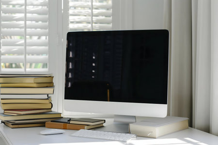 A clean and modern home office setup featuring a computer, stack of books, and natural light from a window.の写真素材