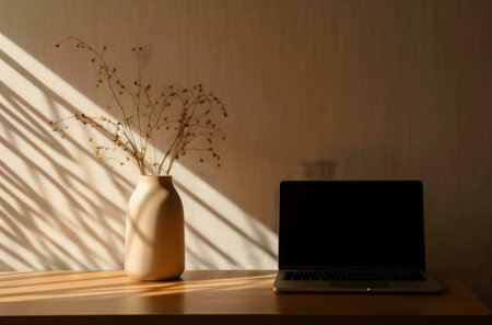 Dried flowers in a vase, next to a laptop on a wooden table. Sunlight casts shadows.の写真素材