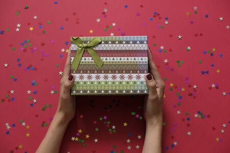 Womans hands holding gift or present box decorated confetti on red table. view from above.