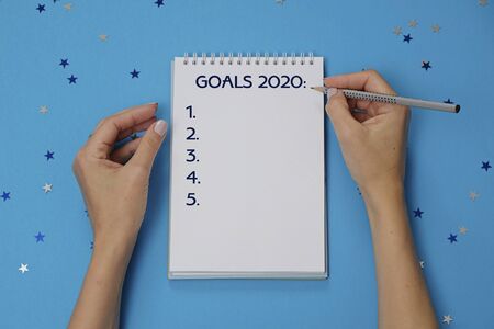 Notebook with text GOALS 2020 and female hands with pencil on festive blue background with stars