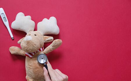 Doctor and christmas concept. Buck get cold, doctor checking him with stethoscope on red background. Place for text.の写真素材