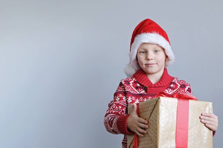 Child in red hat holding Christmas gift box in hand. Boy on gray background. New year and x-mas concept.の写真素材