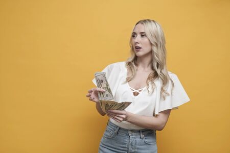 Happy blond woman holding pack of dollars, thinking about shopping or expenses over yellow...