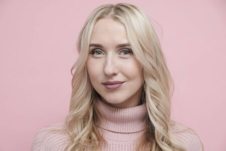 Happy woman face with blond hair in wool sweater on pastel pink background looking at camera. Close...
