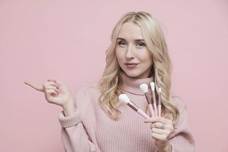 Happy woman face with blond curly hair in wool sweater holding cosmetics brushes on pastel pink background. Close upの写真素材
