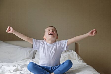 Good morning concept: cute happy little boy stretching and yawning on white bed in a bedroomの写真素材