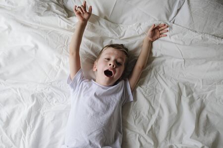 Cute little boy toddler wakes up, yawning and stretching on a bed in a bedroom. Close up, top viewの写真素材