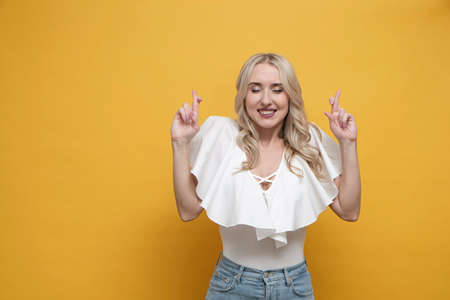 Happy blond european woman in white blouse holding fingers crossed over yellow background. Copy spaceの写真素材