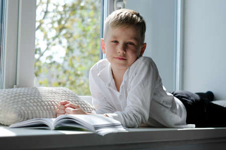 Cute school boy reading a book lying on a windowsill in the house enjoying sunny weatherの写真素材