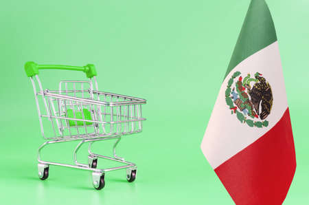 Empty metal shopping cart and flag of Mexico on a colored background, the concept of the consumer...